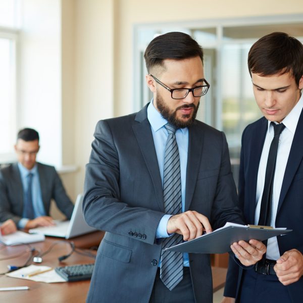 Confident businessman pointing at document while explaining his idea to his partner on background of their colleagues